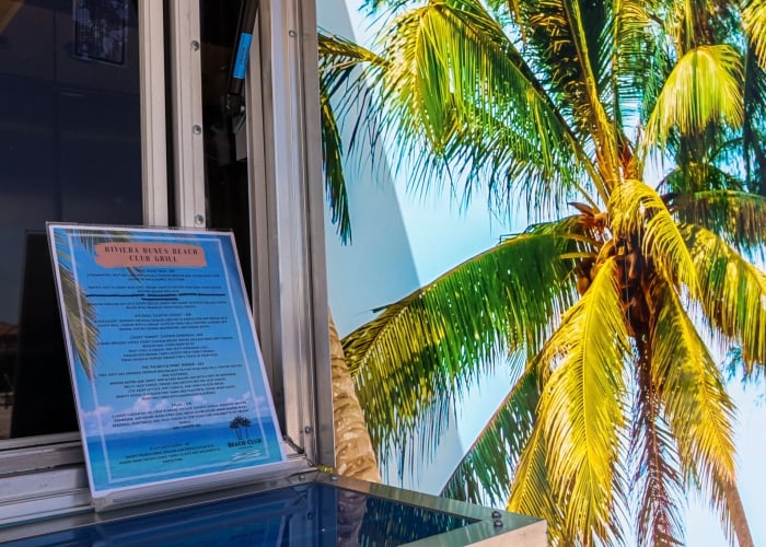 A clear menu for Riviera Dunes Beach Club Grill is displayed next to a vibrant mural featuring a sunny palm tree against a blue sky.