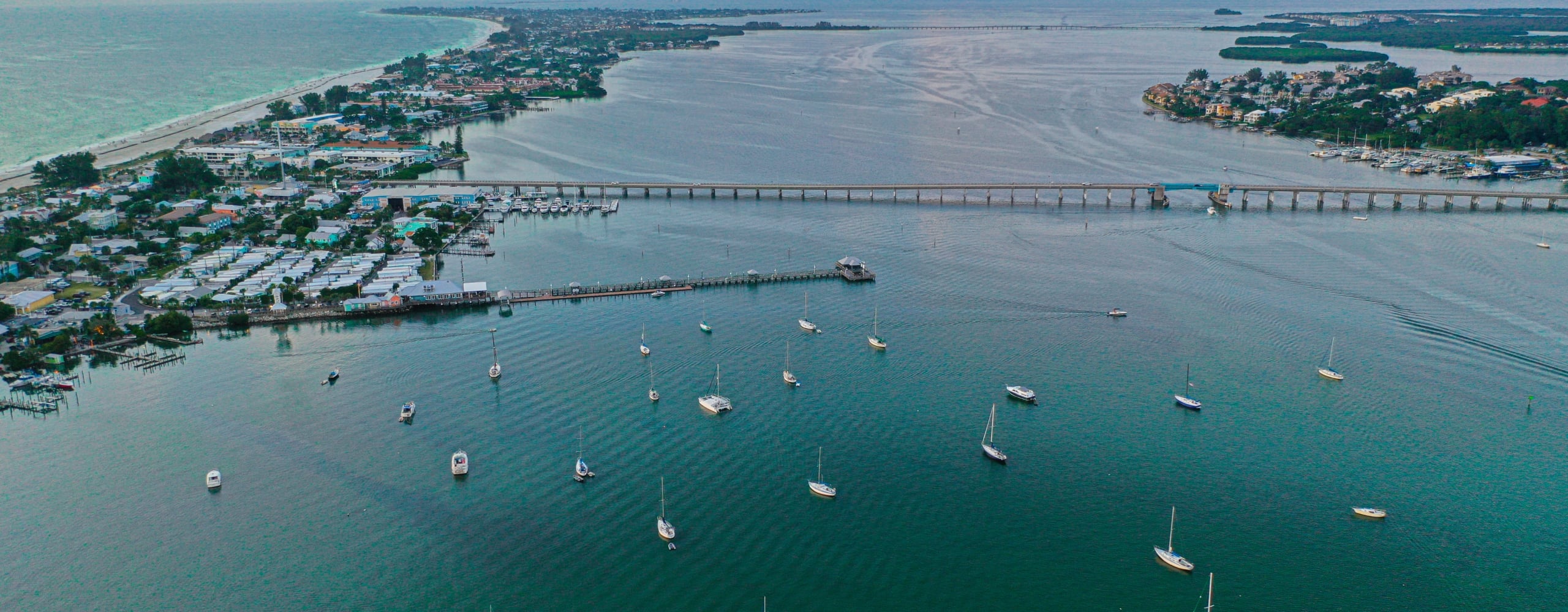 Aerial view of a coastal town with a bridge spanning turquoise water dotted with numerous sailboats near sunset.