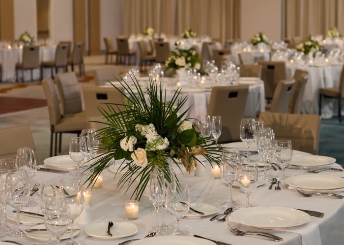 A formally set banquet table with white linens, crystal glassware, and a centerpiece of white roses and green foliage in a softly lit ballroom.