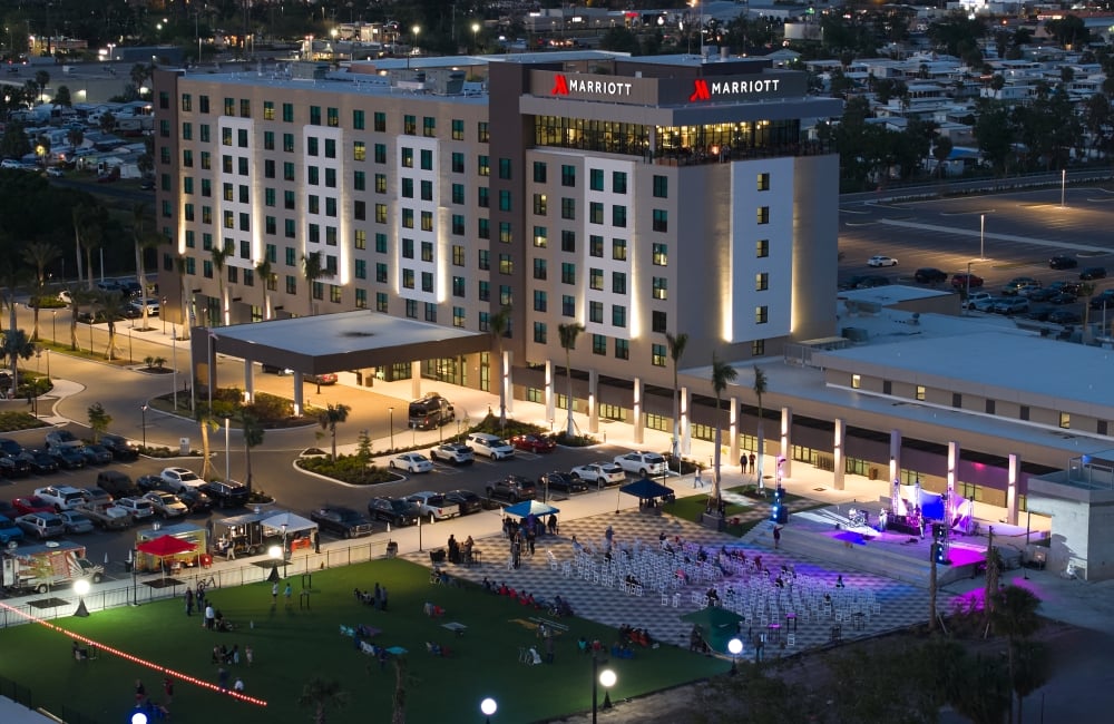 Exterior nighttime view of a brightly lit Marriott hotel building overlooking an outdoor event with a stage, seating, and food vendors on a grassy area.