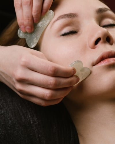 Close-up of a person receiving a facial gua sha massage with two light-colored stones near their forehead and cheek.