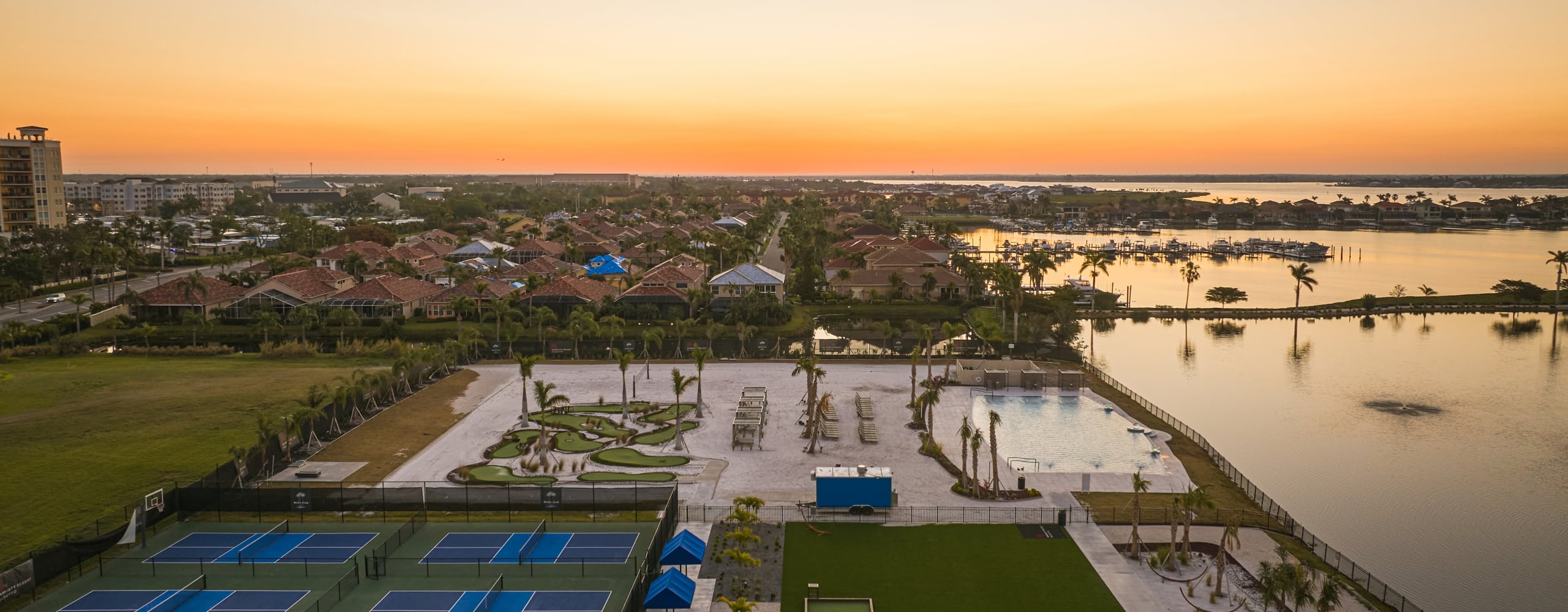 Aerial view of pickleball courts, a pool, and waterfront amenities beside a residential area during an orange sunset.