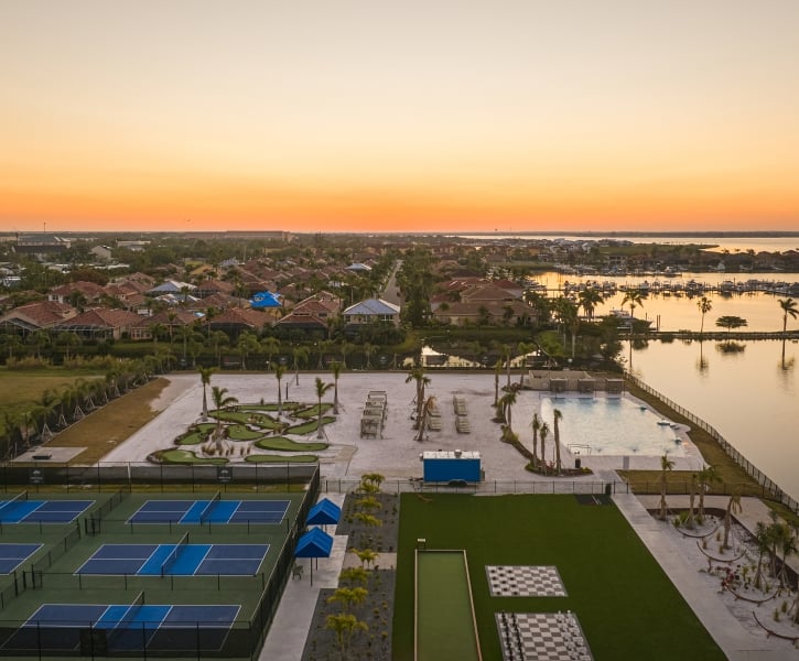 Aerial view of pickleball courts, a pool, and waterfront amenities beside a residential area during an orange sunset.