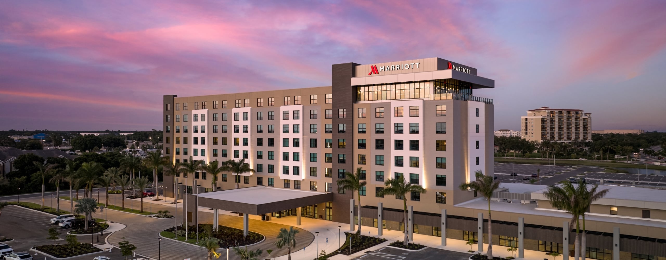 Exterior view of a modern Marriott hotel building at twilight with a vibrant pink and purple sunset sky and surrounding palm trees.