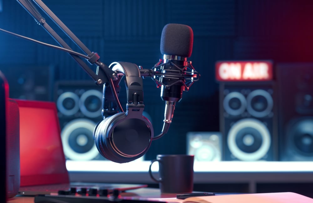 Studio microphone setup with professional headphones hanging nearby, set against a backdrop featuring an "ON AIR" light and studio monitors.