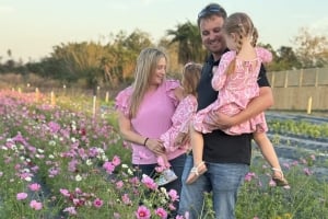 A family standing in a field of blooming pink cosmos flowers at Ananda Farm, with a mother smiling at her two young daughters held in their father's arms.