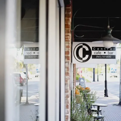 Sign for Central cafe bar hangs outside a brick building on a sidewalk with outdoor seating and a vintage truck parked nearby.