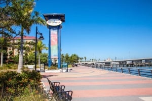 A scenic view of the paved Bradenton Riverwalk promenade on a clear day, featuring the blue Bradenton Riverwalk entrance sign structure, palm trees, park benches, and a bridge over the water in the background.