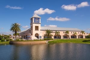 Wide-angle shot of the Ellenton Premium Outlets exterior under a partly cloudy sky, featuring the outlet complex's central tower with its logo, a manicured lawn with palm trees, and a calm pond in the foreground reflecting the scene.
