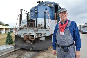 A smiling conductor in striped overalls and a train cap standing in front of a blue and white Florida Railroad Museum locomotive.