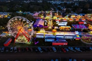 High-angle aerial view of the Manatee County Fairgrounds at night, featuring a brightly lit Ferris wheel, colorful carnival rides, game booths, and rows of parked cars.