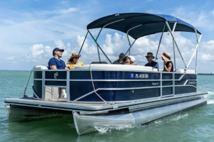A group of people riding on a motorized pontoon boat with a blue canopy out on calm, light-green ocean water under a clear blue sky.