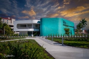 Exterior view of the modern Herrig Center for the Arts building under a dramatic sunset sky, featuring a curved turquoise facade, palm trees, and a paved walkway.