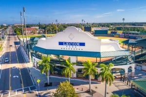 High-angle aerial view of the main entrance and white facade of LECOM Park baseball stadium in Bradenton, showing the adjacent empty road and palm trees lining the entrance under a clear blue sky.