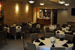 Interior view of the O'Bricks Irish Pub & Martini Bar dining room, featuring an exposed brick wall, multiple round dining tables set with white tablecloths and black napkins, wooden chairs, and warm hanging pendant lights.