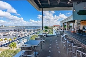 High-angle view of the outdoor, covered rooftop patio and bar area at Oak & Stone, featuring a long bar counter with stools on the right, dining tables and chairs on the left along a glass railing, and a view of a marina with docked boats and the waterfront in the background.