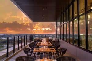 An outdoor, covered rooftop patio dining area at dusk or sunset, featuring a long row of set wooden dining tables and woven chairs along a glass railing that overlooks a distant city skyline under a dramatic orange and cloudy sky.