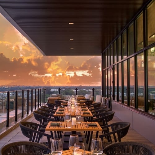 An outdoor, covered rooftop patio dining area at dusk or sunset, featuring a long row of set wooden dining tables and woven chairs along a glass railing that overlooks a distant city skyline under a dramatic orange and cloudy sky.