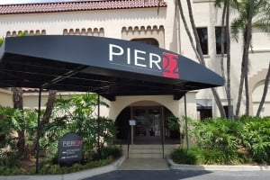 Exterior view of the entrance to the Pier 22 restaurant, featuring a large black awning with the "PIER 22" logo, a smaller directional sign on the left for the restaurant, patio, and ballroom, and palm trees and lush green landscaping surrounding the building's facade.