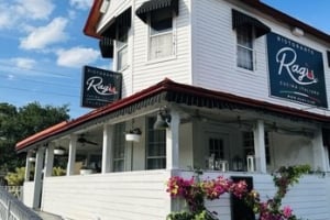 Exterior view of the Ragù Cucina Italiana restaurant, featuring a white wooden multi-story building with a covered porch or patio dining area, outdoor floral arrangements, and prominent dark signs featuring the restaurant's logo.
