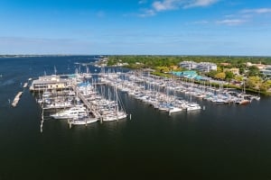 High-angle aerial view of the Regatta Pointe Marina on a clear day, featuring rows of numerous boats and yachts docked along the piers, with buildings and trees lining the waterfront in the background.