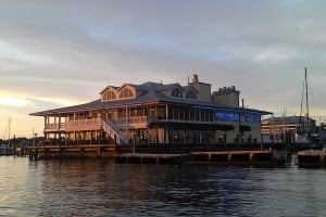 Wide-angle view of the Riverhouse Waterfront Restaurant building situated over the water at dusk, featuring glowing indoor and outdoor lighting, with docked sailboats and a dramatic sunset sky in the background.