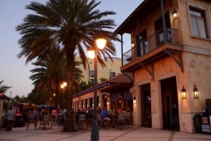 Exterior view of Riviera Dunes Dockside at dusk, featuring a multi-story building with a stucco facade and balconies, illuminated by warm outdoor street lamps and building lights, with a tall palm tree on the left and patrons dining at outdoor tables in the background.