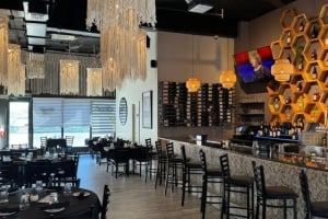 Interior view of the modern Sofra Kitchen Bar & Bistro dining area and bar, featuring black dining tables, a long bar counter with stools, prominent honeycomb-shaped shelving units on the wall, and decorative hanging fringe light fixtures.