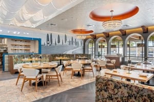 Bright, wide interior shot of a modern restaurant dining room featuring round wooden tables, upholstered chairs, arched windows, patterned booths, and warm, decorative light fixtures.