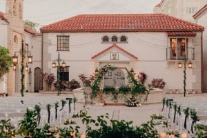 An outdoor courtyard wedding setup at The Bishop Museum of Science and Nature, featuring a stone building with a red-tile roof, a floral-adorned hexagonal arch by a central fountain, and rows of white chairs.