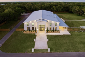 Elevated aerial view of The Elia event venue featuring a modern white barn with a silver roof, illuminated windows, and a central walkway surrounded by green lawns.