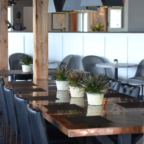 Interior view of a dining room showcasing a long wooden table set with black chairs, place mats, and small potted plants, illuminated by geometric pendant lights overhead.