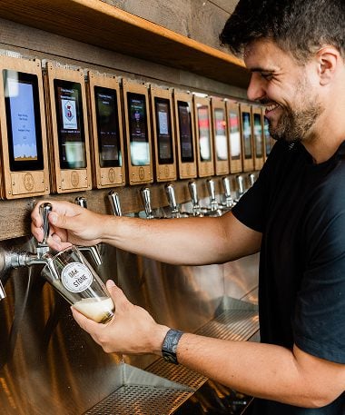 A man smiling as he pours a drink from a self-serve beer tap wall into a branded glass.