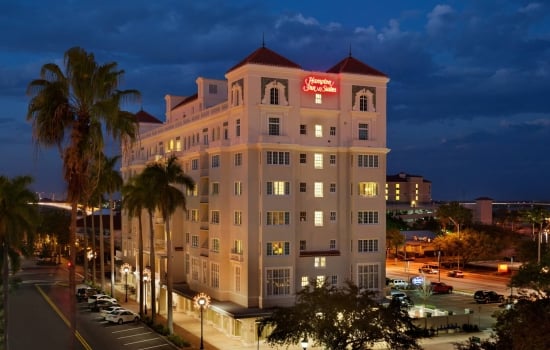 Exterior dusk view of the multi-story Hampton Inn & Suites building illuminated at night, featuring a lit red rooftop sign, a tree-lined street with a parking lot in front, and a deep blue evening sky.