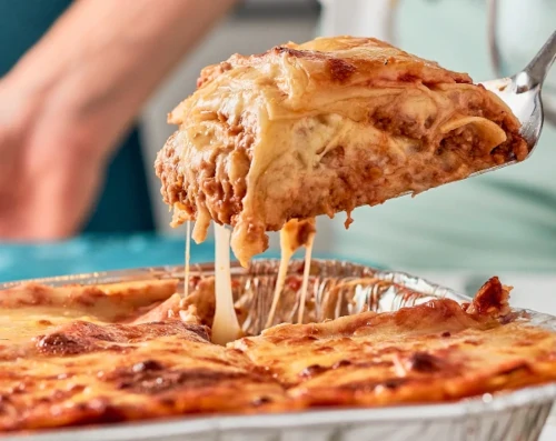 A large slice of multi-layered lasagna being lifted with a metal spatula from a baking pan, showing melted, stretching cheese.