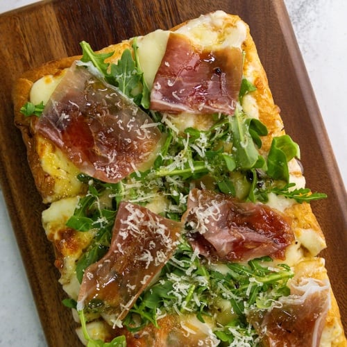 Top-down view of a rectangular flatbread pizza on a wooden serving board, topped with melted cheese, fresh green arugula, slices of prosciutto, and grated parmesan cheese.