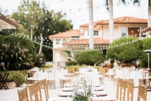 An elegant outdoor wedding reception setup at Palmetto Riverside Bed & Breakfast featuring a long dining table dressed in a white tablecloth, topped with greenery centerpieces and candles, and surrounded by gold chiavari chairs under palm trees and string lights.