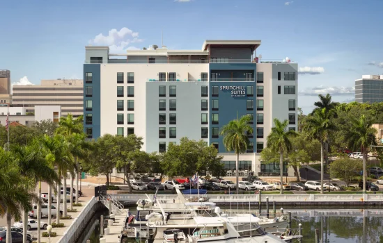 Exterior view of the multi-story SpringHill Suites building situated behind a marina filled with docked boats, flanked by palm trees and a paved walkway under a clear sky.