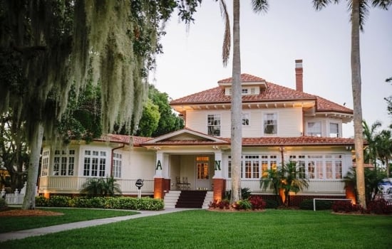 Exterior view of a historic, two-story white inn with a tiled roof, surrounded by a manicured green lawn, tall palm trees, and moss-draped trees.