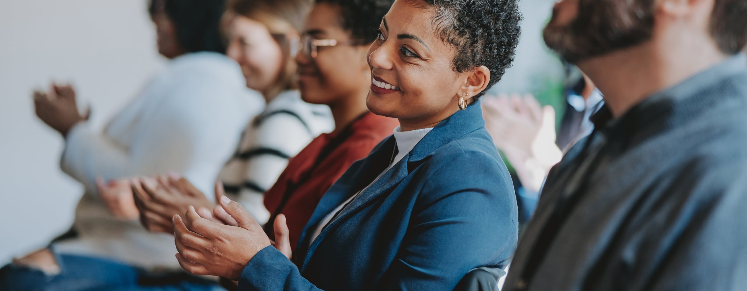 A smiling professional woman and a man with a beard sitting in an audience and clapping during a presentation or conference.