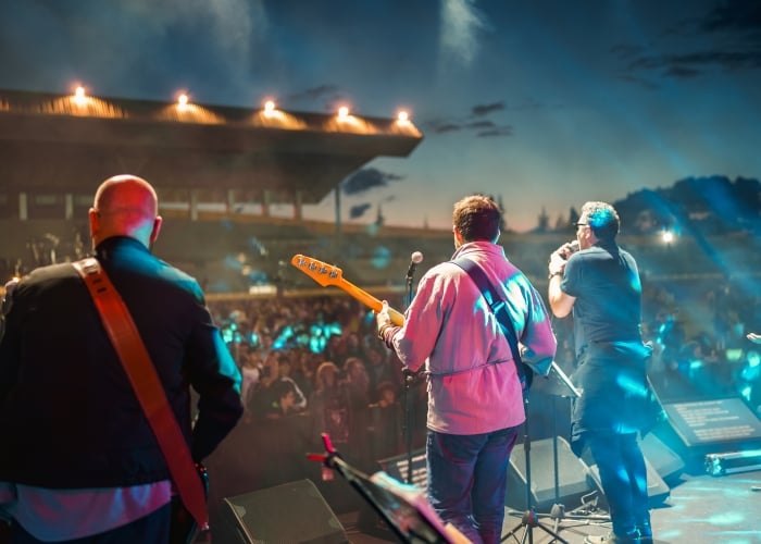 Rear view of a live band performing on an outdoor stage at night before a large crowd, with stage lights and string lighting overhead.
