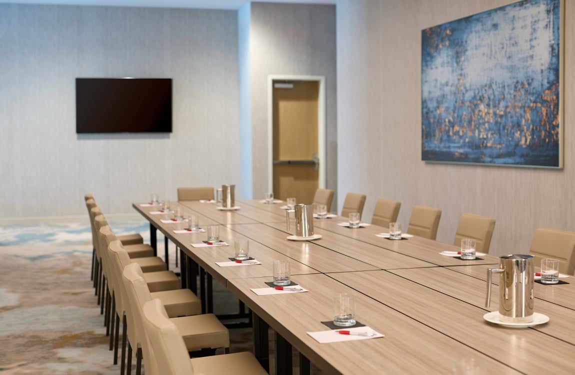 A long wooden conference table set with water glasses, note pads, and tan chairs in a modern meeting room, featuring a large abstract blue painting on the wall and a flat-screen TV.