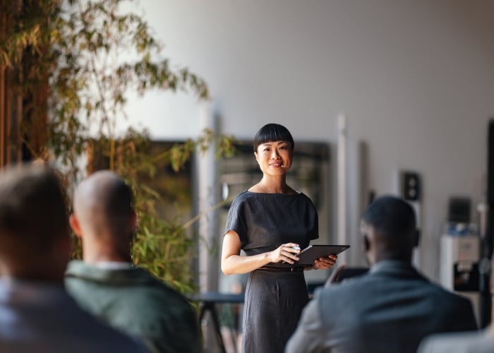A professional woman standing and presenting to a group of seated colleagues during a business meeting or workshop, holding a digital tablet.