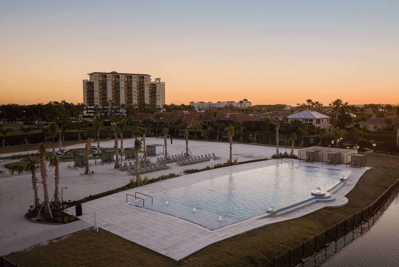 Elevated aerial view of a large, modern resort swimming pool at dusk, with a sandy beach lounging area, lounge chairs, palm trees, and the multi-story resort building standing in the background under a sunset sky.
