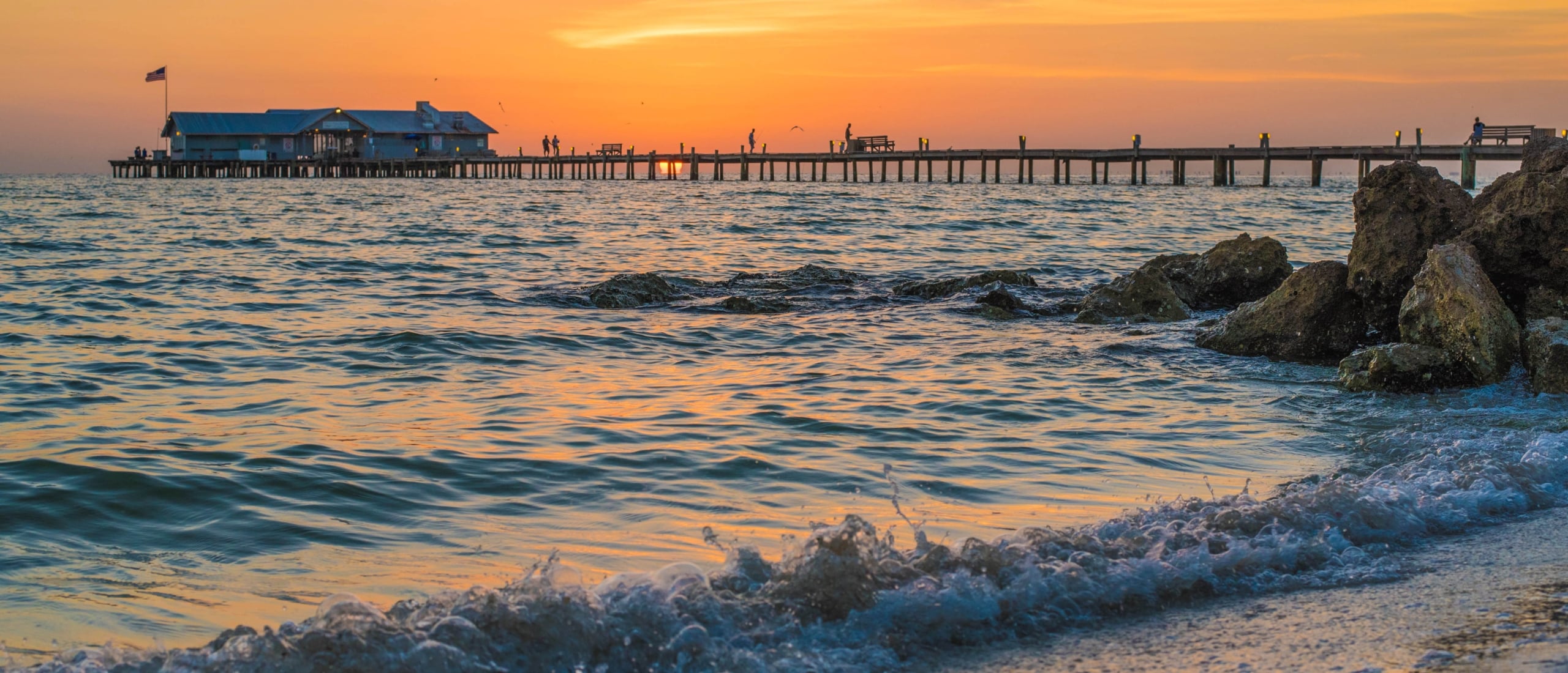 A vibrant sunset view from a sandy beach looking toward the Rod and Reel Pier in Anna Maria Island, with waves crashing on the shore and rocks in the foreground.