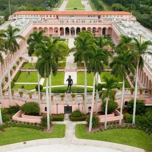 High-angle view of the pink, Italianate courtyard at The John and Mable Ringling Museum of Art featuring tall palm trees, statues, and manicured lawns.