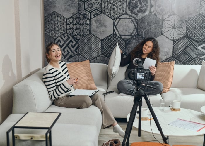 Two smiling women in a modern studio space with patterned wallpaper, filming content with a camera set up on a tripod in front of a white sofa.