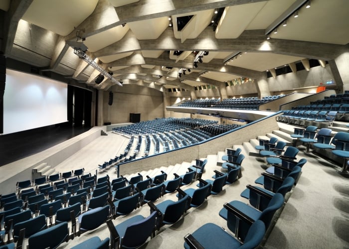 A high-angle interior view of a large auditorium or theater featuring curved rows of blue stadium seating facing a large projection screen on the left.